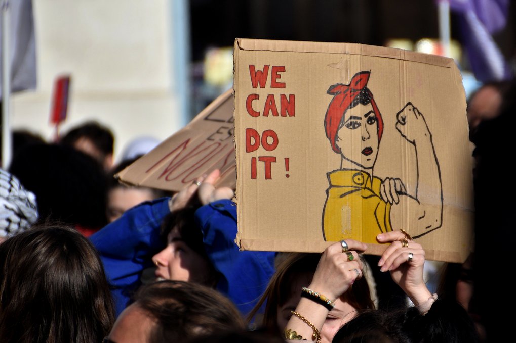 A demonstration at International Women's Day, women are seen holding up a cardboard sign with the slogan  "We Can Do It!" and the illustration of a woman flexing her arm, symbolizing empowerment.