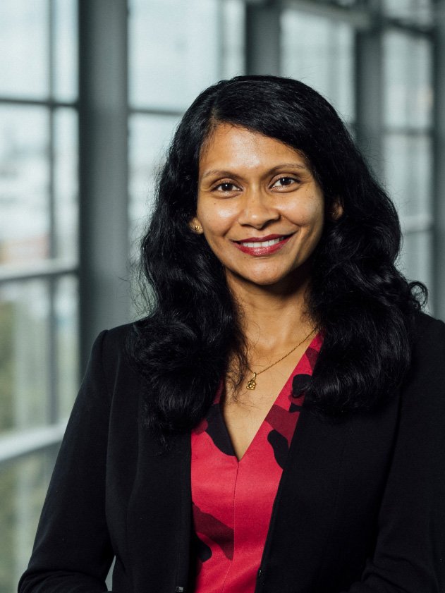 A woman wears a black blazer over a red patterned top. Large windows with natural daylight are visible in the background.