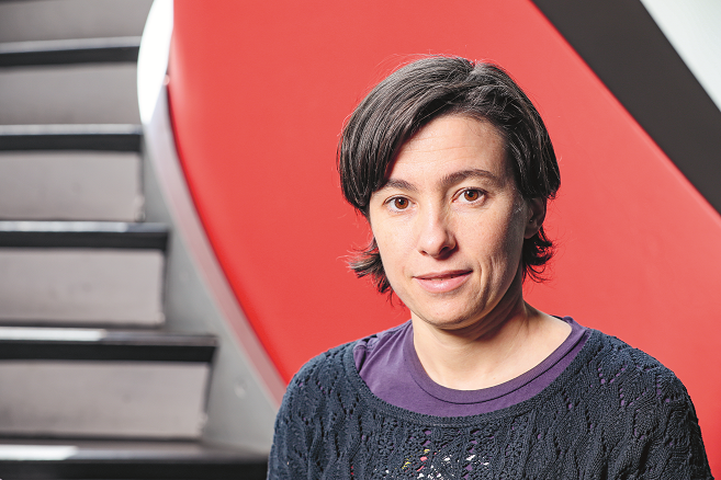 Portrait of Carmela Troncoso (smiling, dark hair parted in the middle) in front of a winding staircase with red railings.