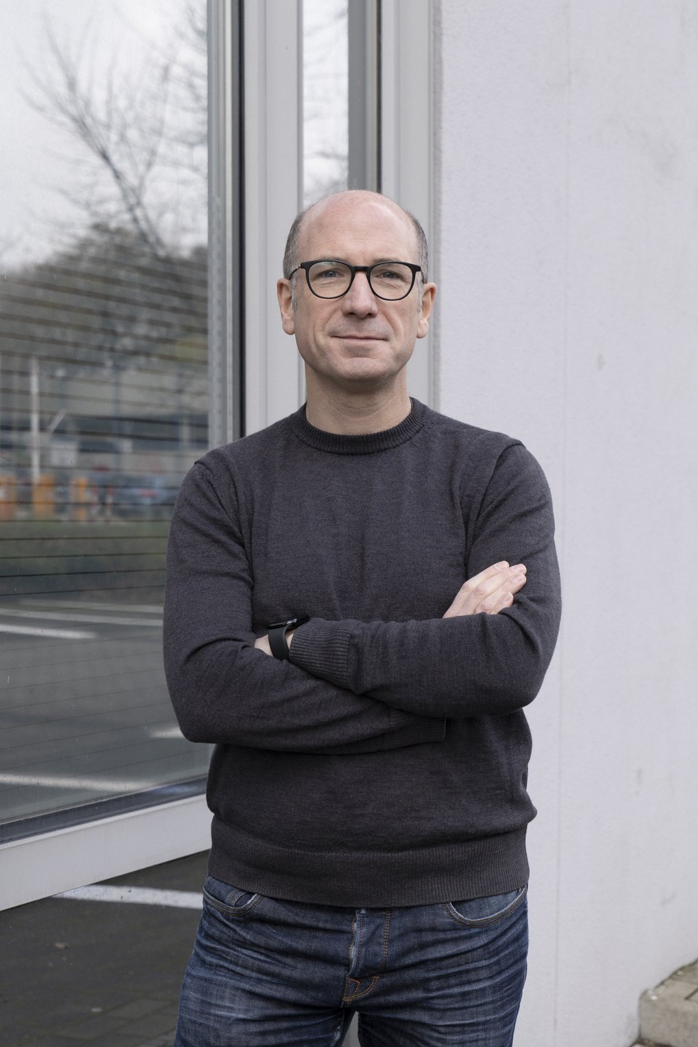 Ein Mann mit Brille und dunklem Pullover steht mit verschränkten Armen vor einem Fenster, in dem sich Bäume und ein Parkplatz spiegeln.