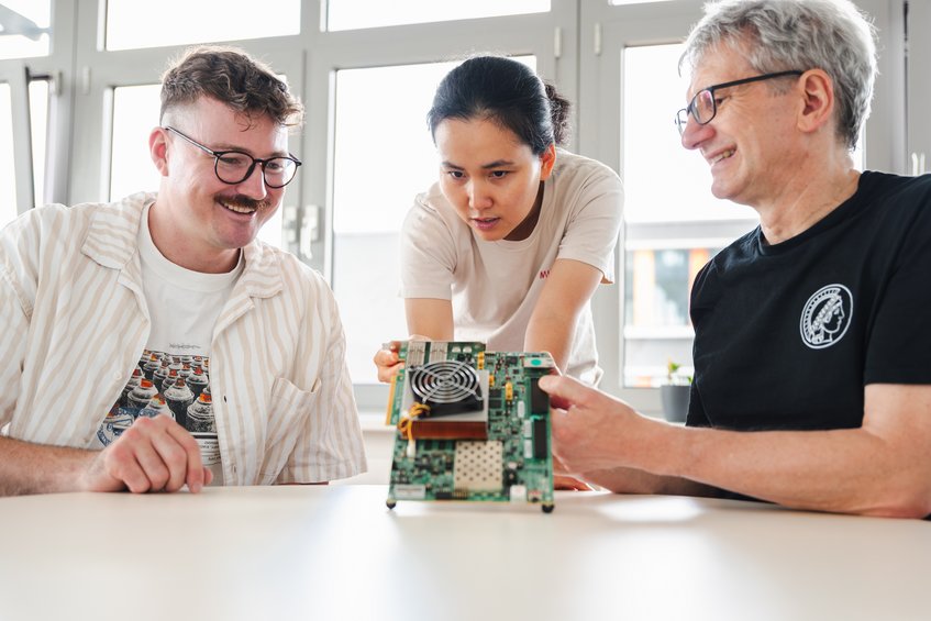 Komplizierte Technik: Julian Speith, Hoang Nguyen Hien Pham und Christof Paar (von links) begutachten eine Platine, auf der Mikrocontroller und andere elektronische Bauteile derart viel Wärme erzeugen, dass ein großer Lüfter auf der Oberseite diese aktiv kühlen muss. Die Forschenden Julian Speith, Hoang Nguyen Hien Pham und Christof Paar schauen sich eine Platine an.