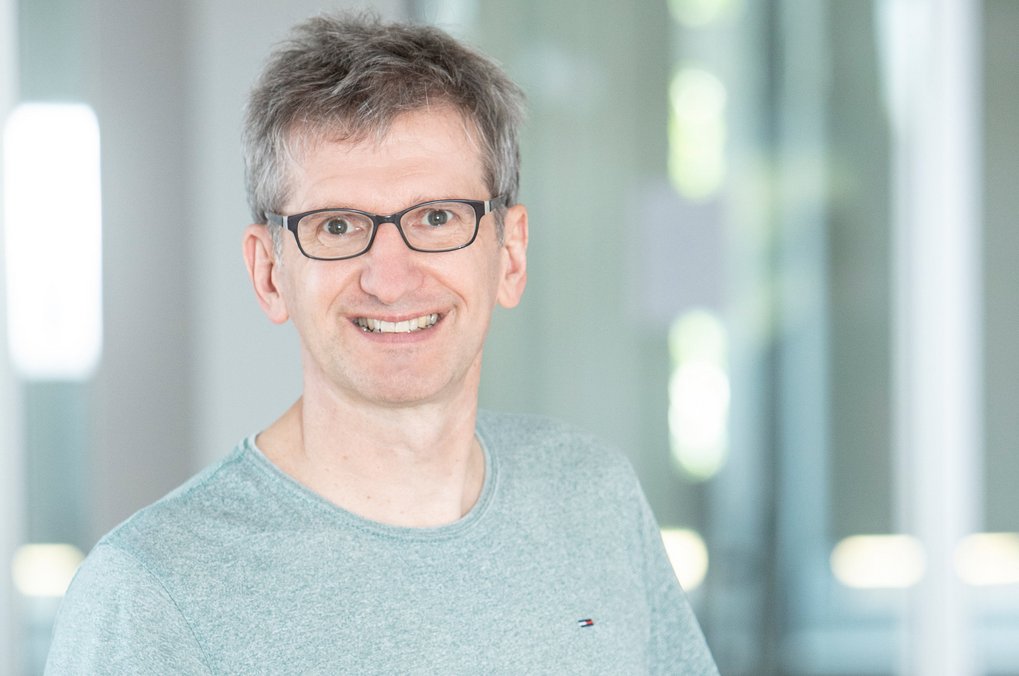 Mann mit Brille und grauem T-Shirt vor unscharfem Hintergrund.