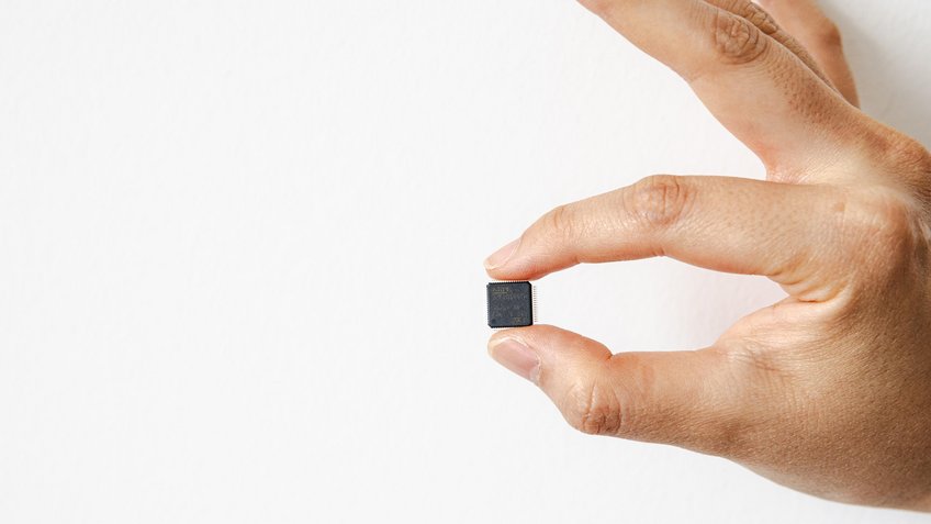Barely larger than a fingernail: This chip is like a tiny computer. Sixty-four metal pins protrude from the black casing. Some of these contacts are used for power, for example, but the vast majority are used for the input and output of commands and signals. A hand is holding a small black microchip between its thumb and index finger.