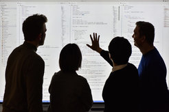 Vier Personen, die nur umrissartik zu erkennen sind, betrachten eine Projektion von Software-Code auf eine Leinwand.