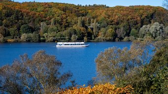 Autumn at Kemnader See, a nearby lake. A white boat on a blue lake surrounded by trees in autumnal colours.